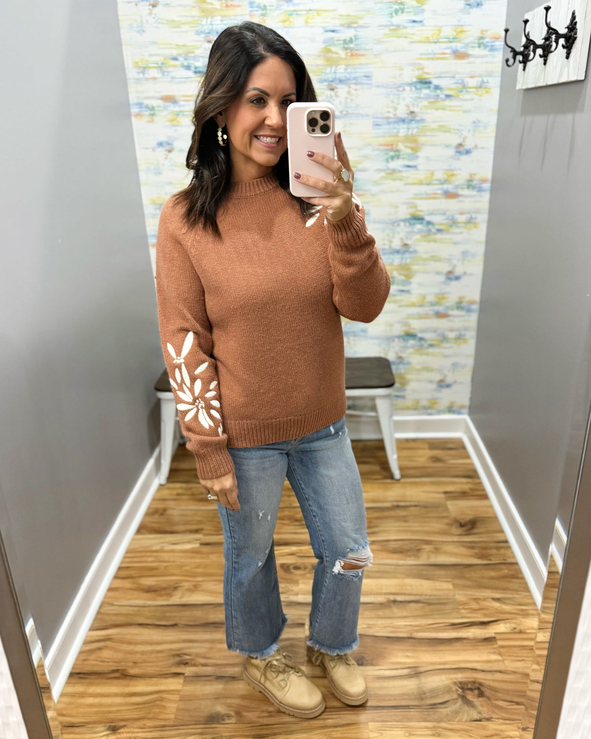 A woman takes a mirror selfie in a fitting room wearing a rust-colored knit sweater with white floral embroidery on one sleeve, paired with distressed blue jeans and beige boots.