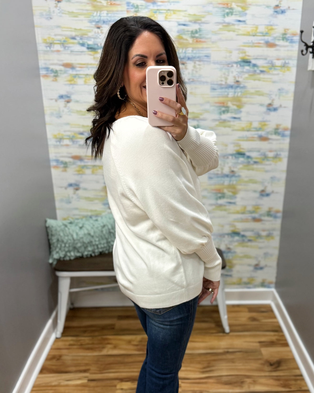 A woman takes a mirror selfie wearing a cream-colored knit sweater and dark blue jeans in a room with patterned wallpaper and a wooden floor.