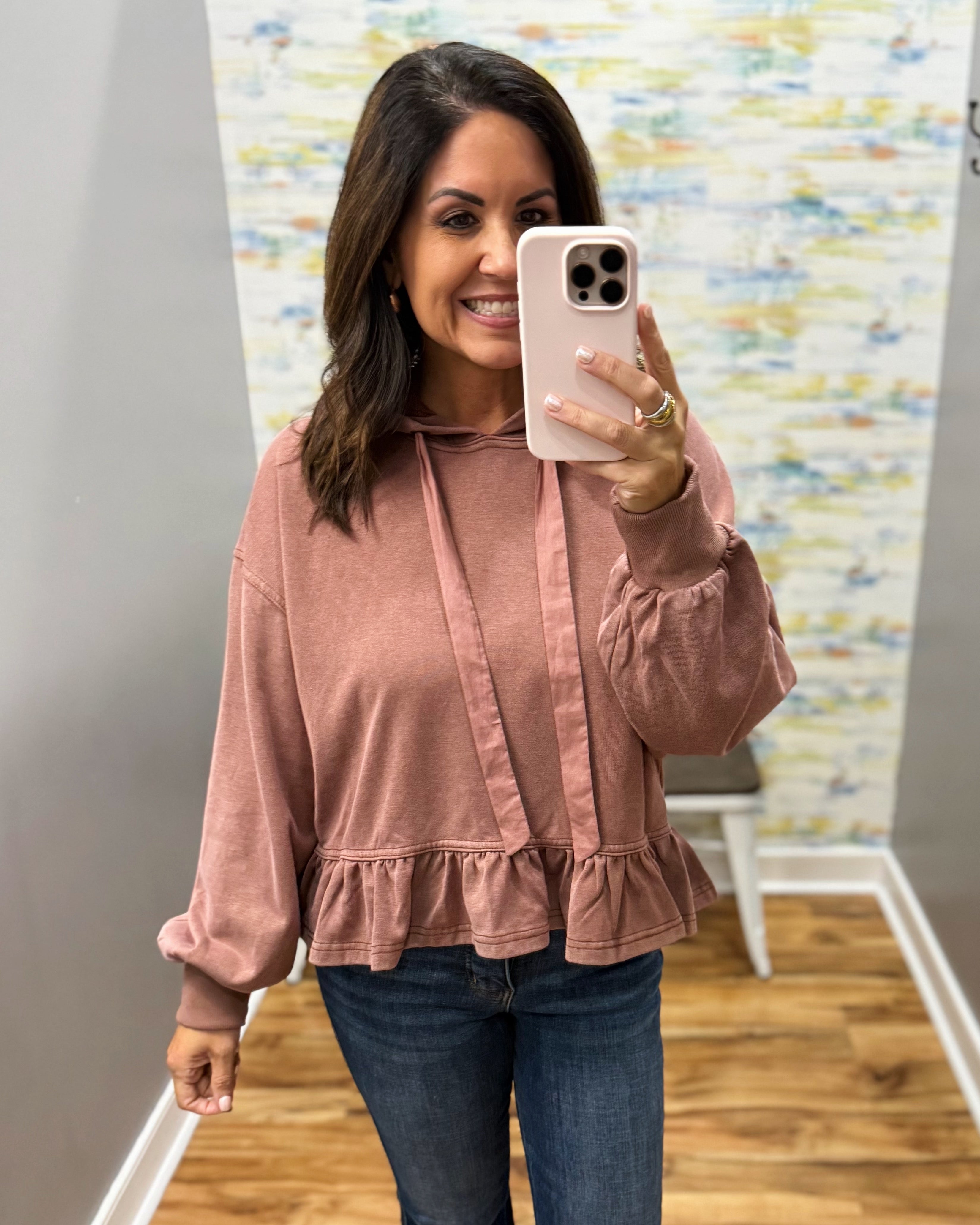 A woman taking a mirror selfie wearing a dusty rose long-sleeve hoodie with a ruffled hem and tie detail, paired with blue jeans.