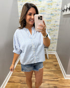 Woman taking a mirror selfie wearing a blue and white striped shirt and denim shorts.