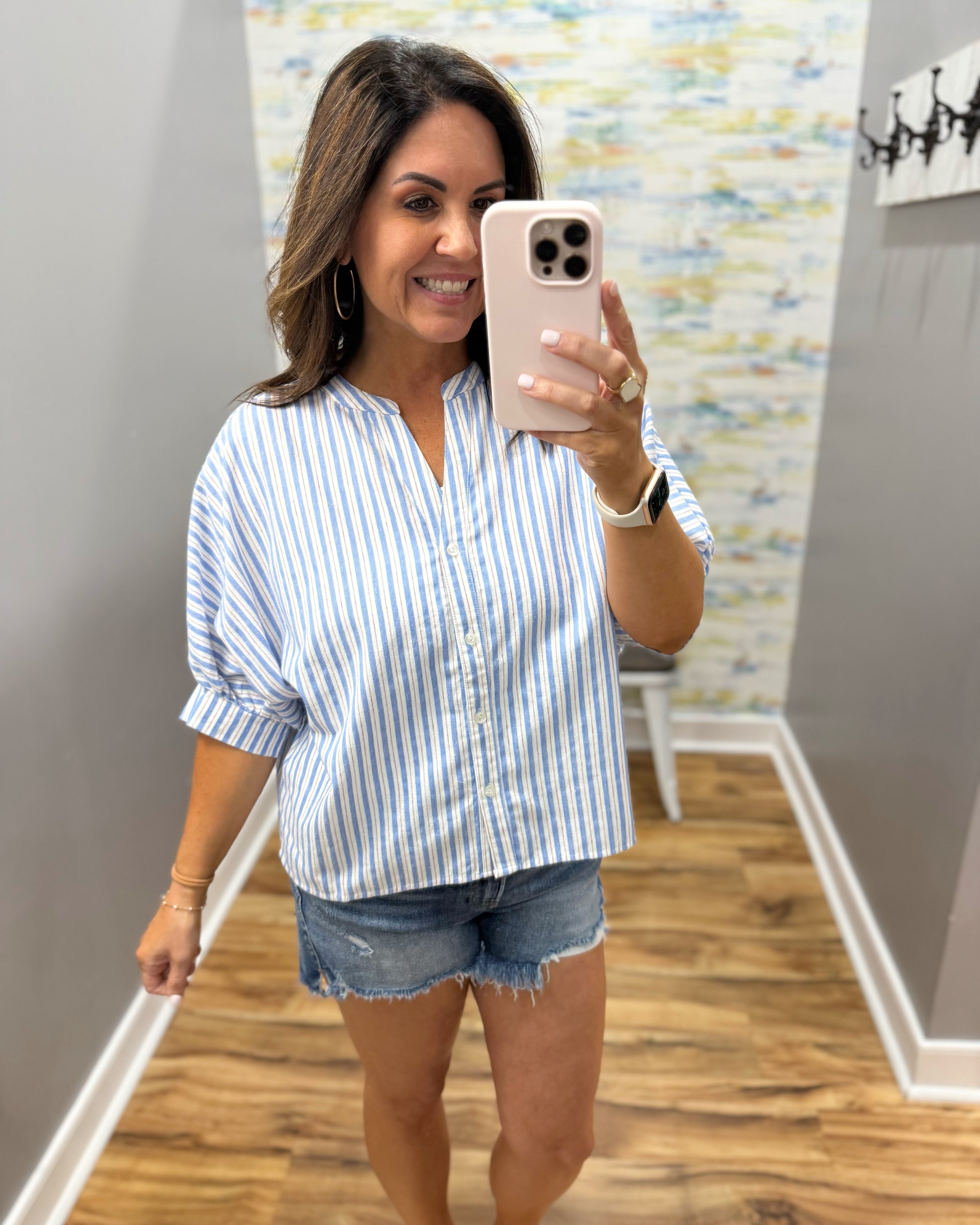 Woman taking a mirror selfie wearing a blue and white striped shirt and denim shorts.