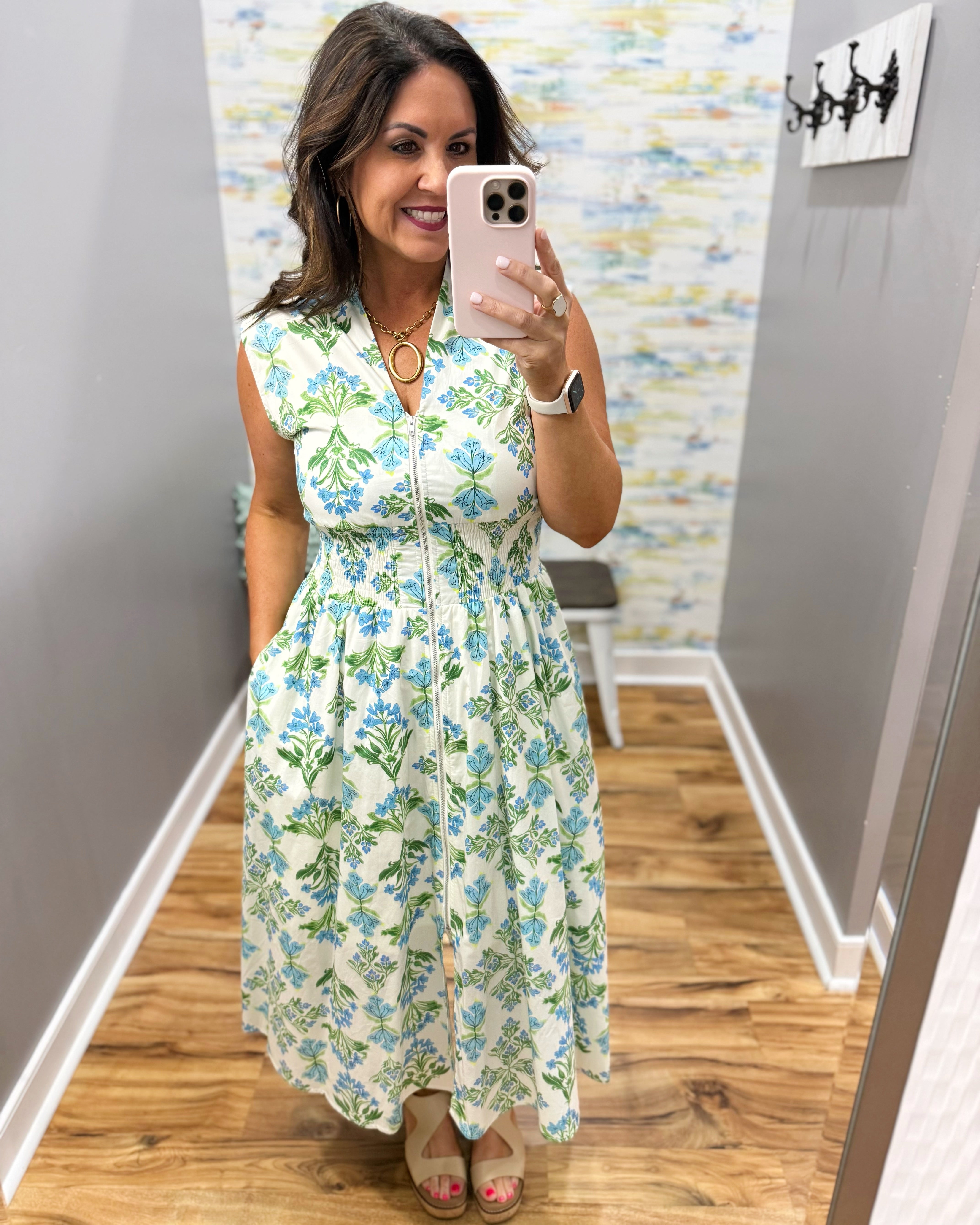 Woman taking a mirror selfie wearing a floral dress in a room with patterned wallpaper.