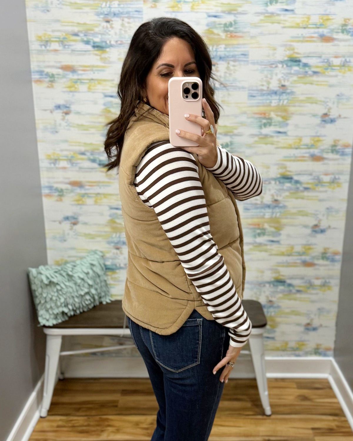 A woman takes a mirror selfie wearing a tan corduroy puffer vest over a brown and white striped long-sleeve shirt and dark blue jeans. The background shows a gray wall, a white bench with a mint green pillow, and a colorful abstract wallpaper.