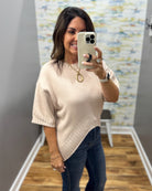 A woman takes a mirror selfie wearing a beige knit sweater with a diagonal ribbed hem and dark blue jeans in a fitting room.