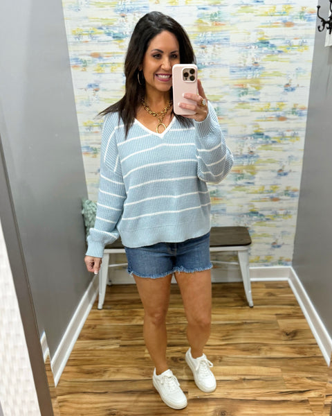 Woman wearing a light blue striped sweater, denim shorts, and white sneakers in a room with patterned wallpaper.