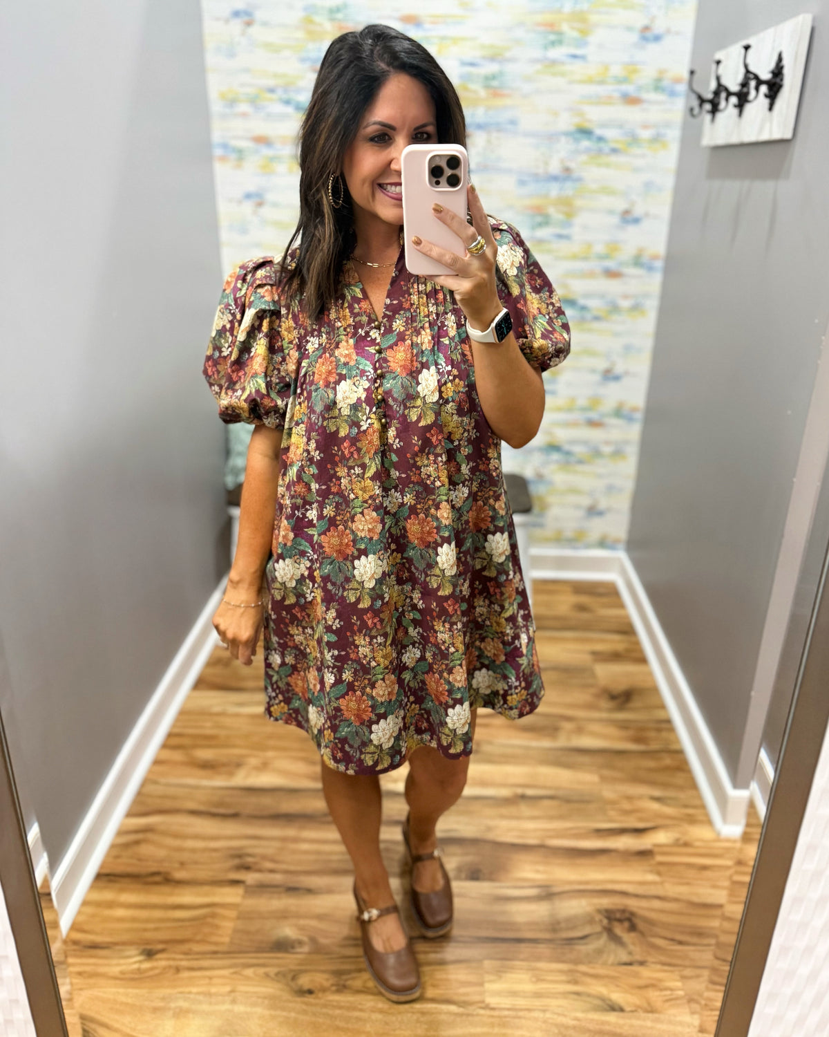 A woman takes a mirror selfie in a fitting room wearing a short puff-sleeve floral dress with a burgundy base and multicolor flowers, paired with brown flat shoes.