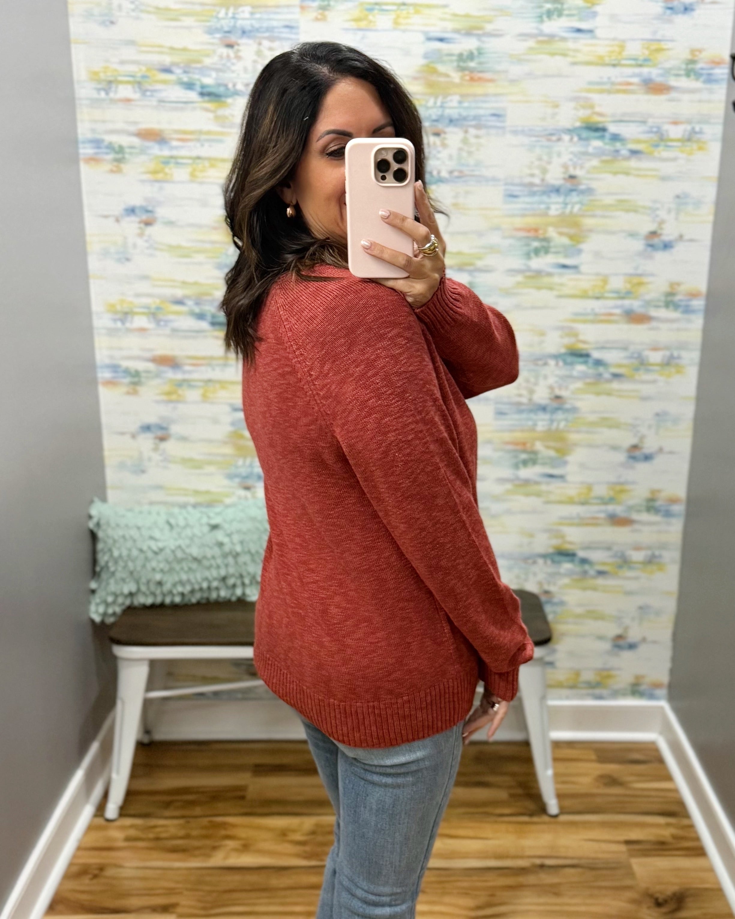 A woman takes a mirror selfie wearing a heathered rust-red knit sweater and light-wash blue jeans in front of a patterned wallpapered wall.