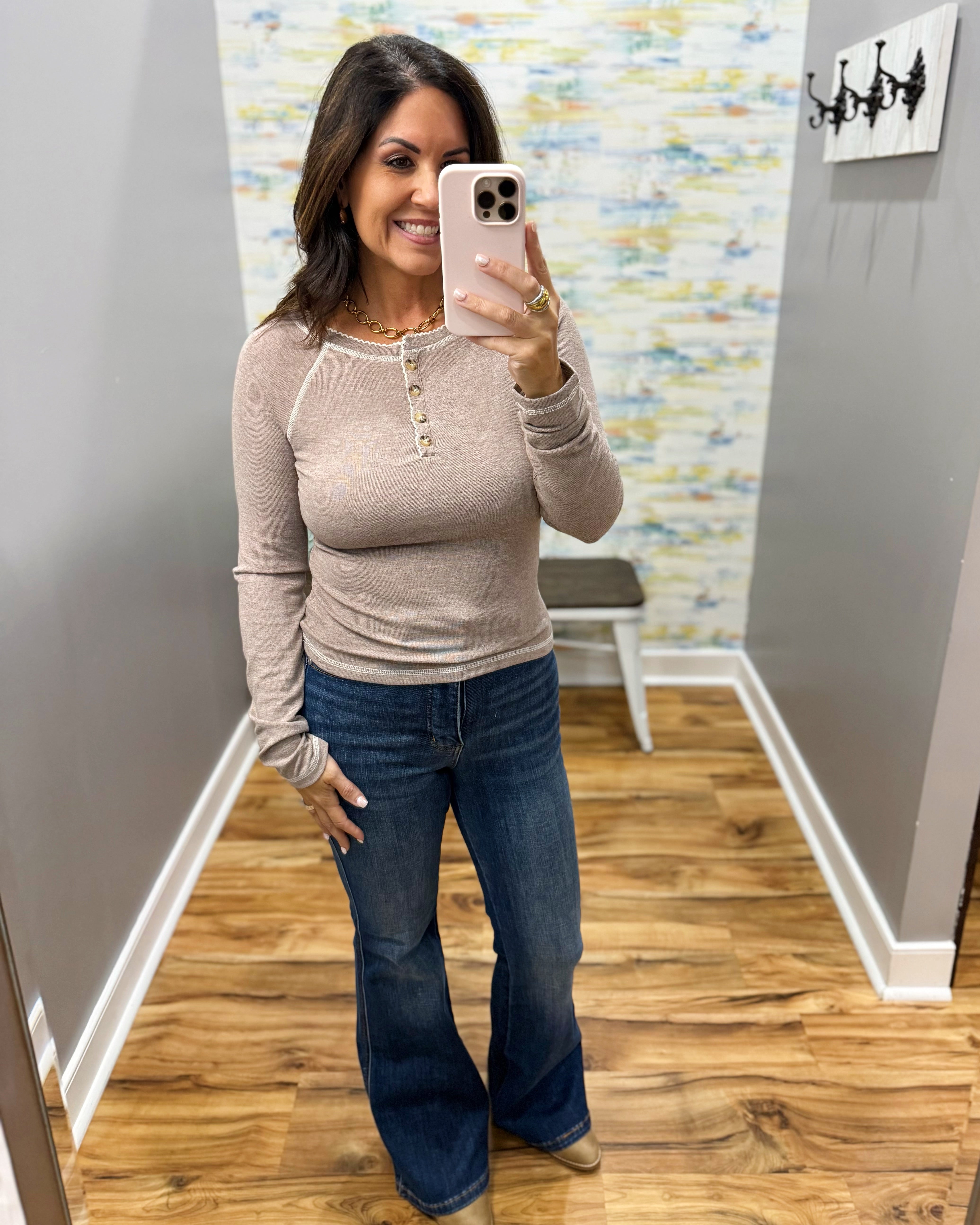 A woman takes a mirror selfie in a fitting room wearing a beige long-sleeve henley top and blue flare jeans.