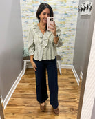 A woman takes a mirror selfie in a fitting room wearing a light sage green and white vertically striped ruffled blouse with elbow-length sleeves, paired with dark navy wide-leg trousers and brown shoes.