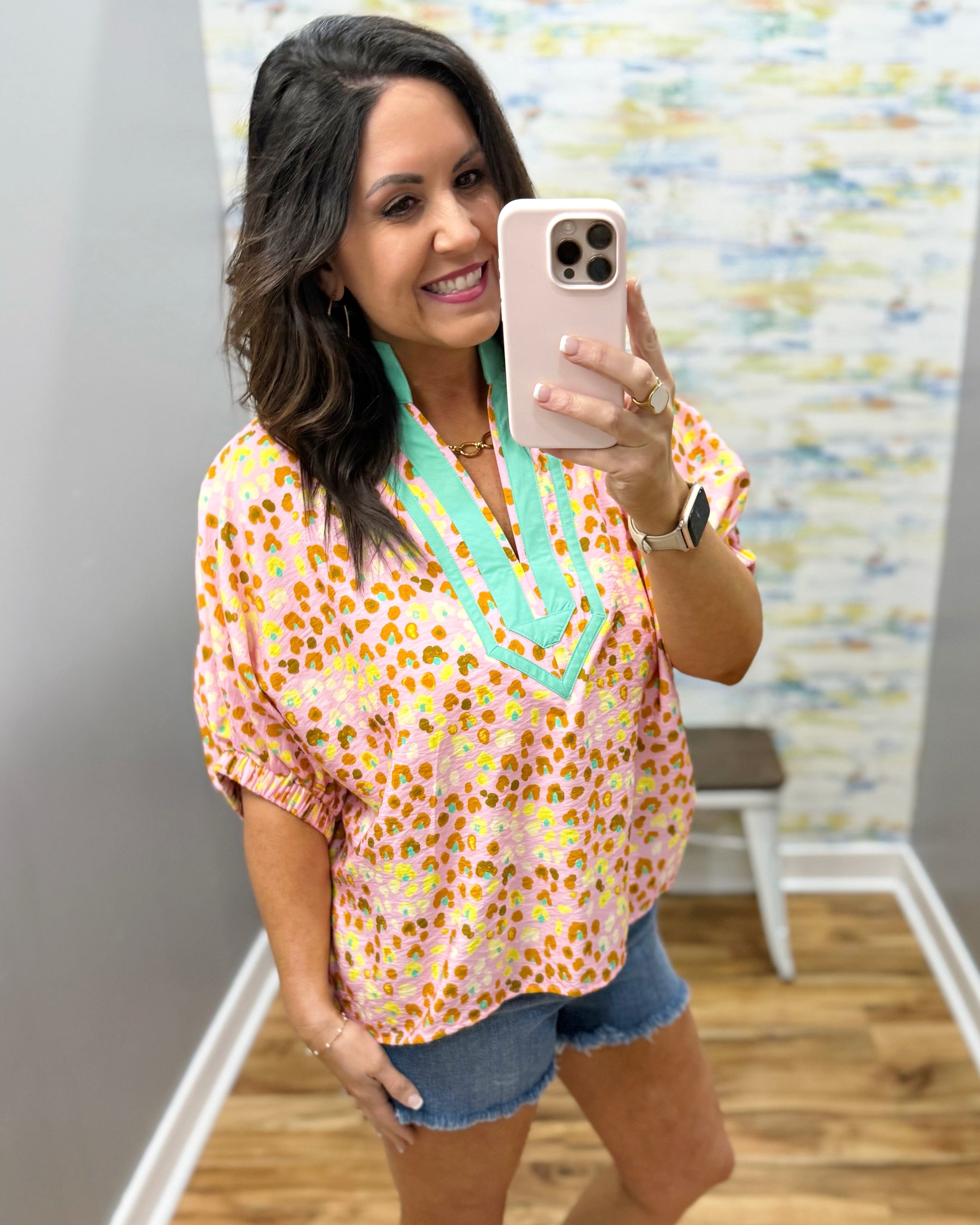 Woman taking a mirror selfie wearing a floral blouse with green trim and denim shorts.
