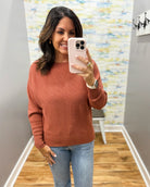 A woman takes a mirror selfie wearing a textured, long-sleeve crewneck sweater in a muted terracotta or burnt orange color, paired with light-wash blue jeans.