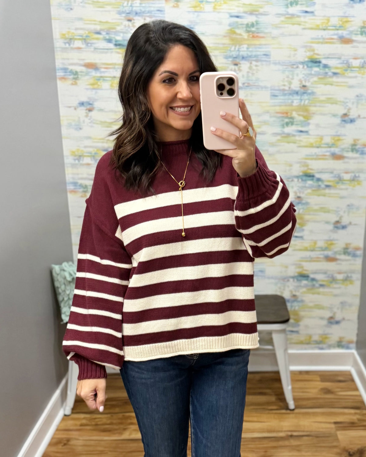 A woman takes a mirror selfie wearing a burgundy and cream striped knit sweater, blue jeans, and a gold necklace in a room with patterned wallpaper.
