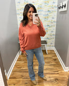 A woman takes a mirror selfie wearing a terracotta orange long-sleeve V-neck sweater, blue jeans, and tan shoes in a home hallway with wood floors and gray walls.