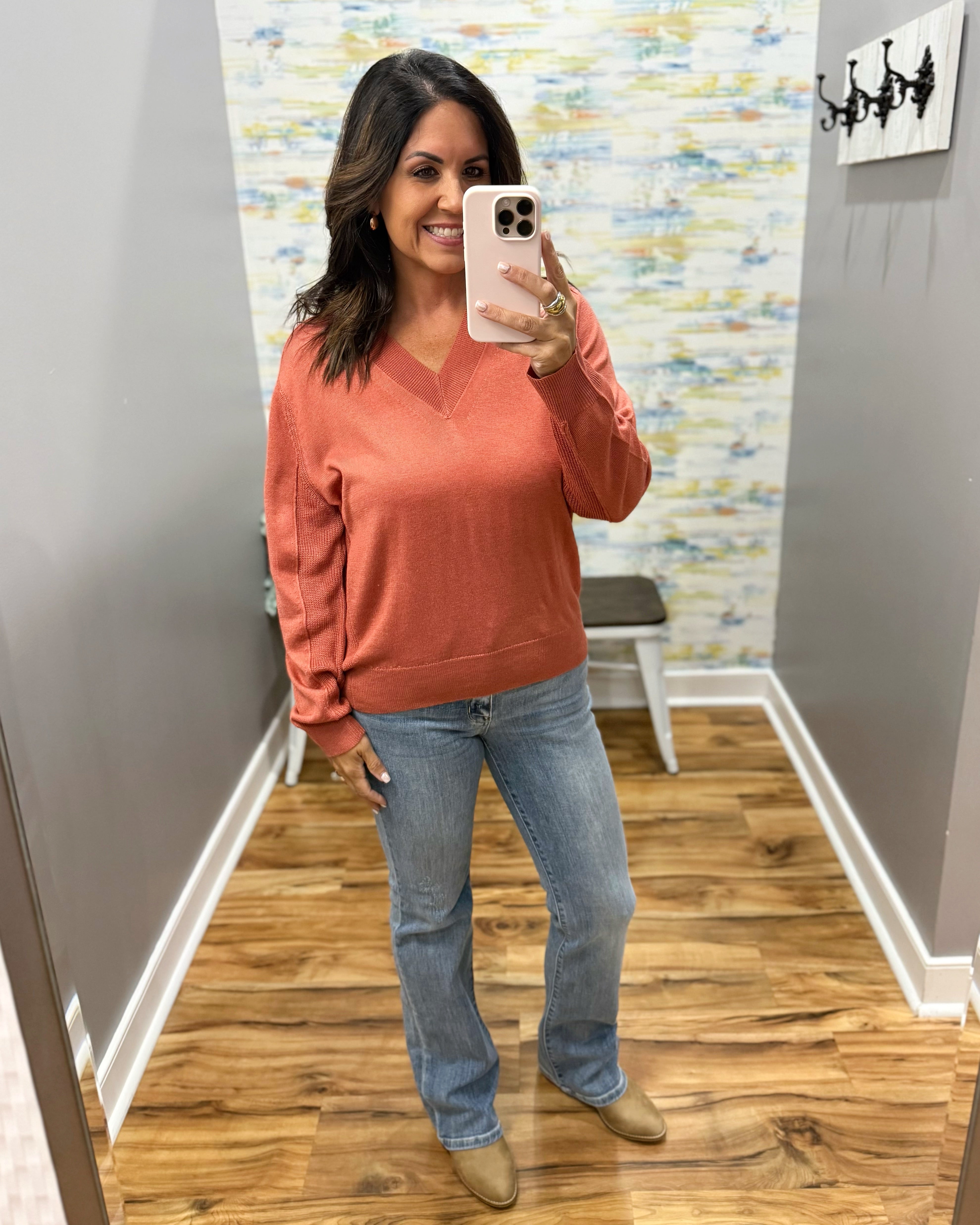 A woman takes a mirror selfie wearing a terracotta orange long-sleeve V-neck sweater, blue jeans, and tan shoes in a home hallway with wood floors and gray walls.
