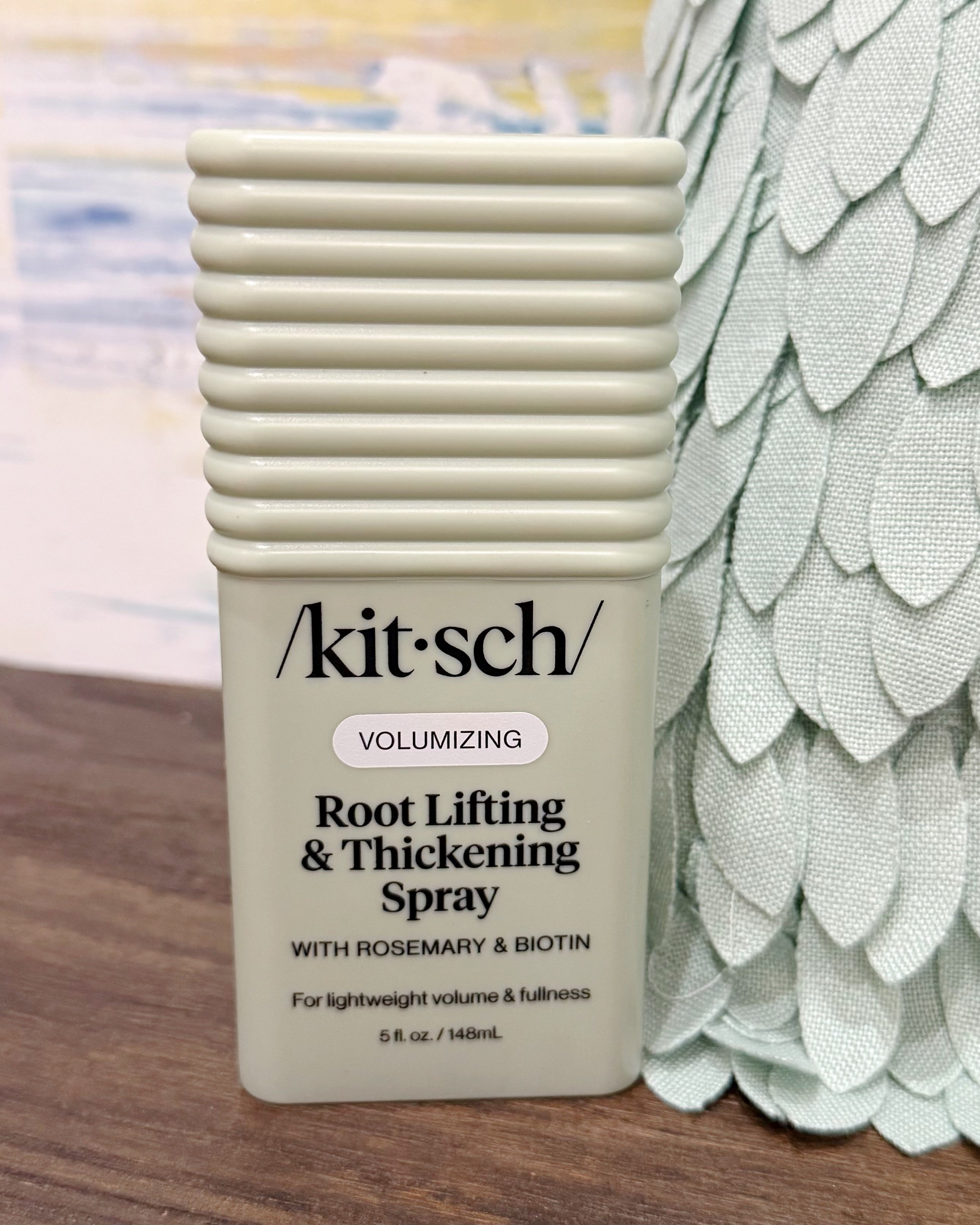 A bottle of Kit-Sch Volumizing Root Lifting & Thickening Spray with rosemary and biotin, shown on a wooden surface next to a decorative green textured prop.
