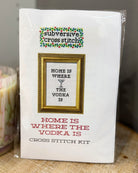 A packaged cross stitch kit labeled 'subversive cross stitch' with a framed design reading 'HOME IS WHERE THE VODKA IS' on the front, photographed standing on a fabric surface.