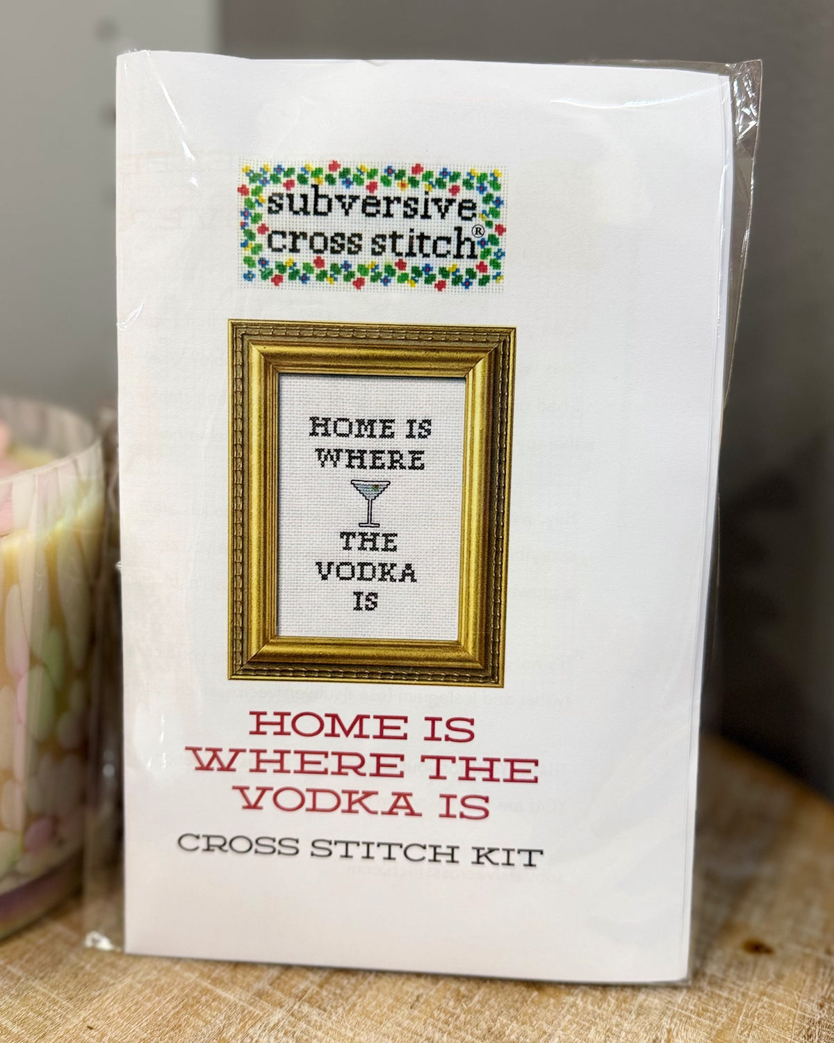 A packaged cross stitch kit labeled 'subversive cross stitch' with a framed design reading 'HOME IS WHERE THE VODKA IS' on the front, photographed standing on a fabric surface.