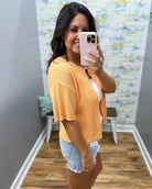 Woman taking a mirror selfie wearing a yellow cardigan and denim shorts in a room with a textured wall.