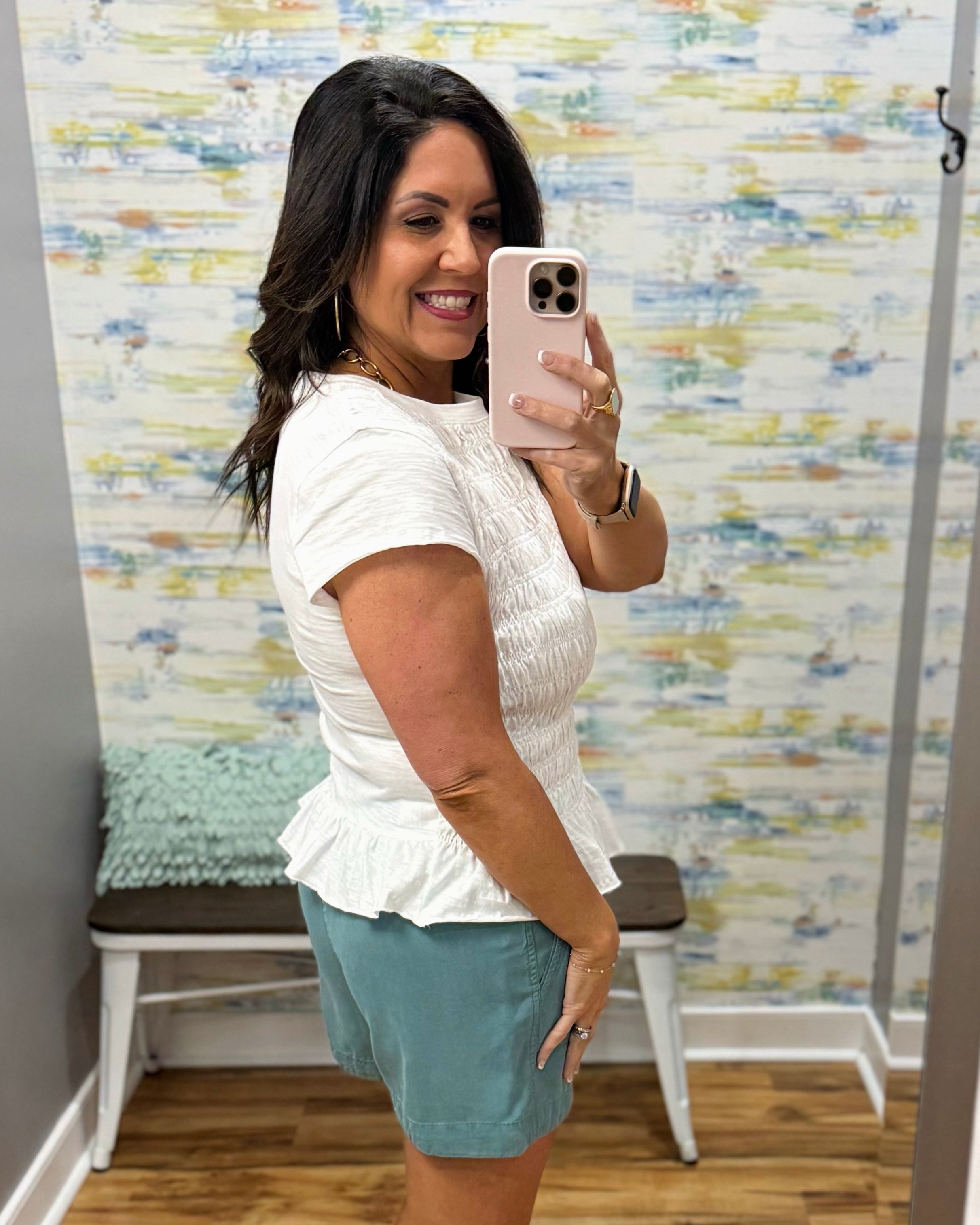 Woman taking a mirror selfie wearing a white top and teal shorts in front of a colorful wall.