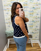 Woman taking a mirror selfie wearing a navy blue sleeveless top with white polka dots and blue jeans.