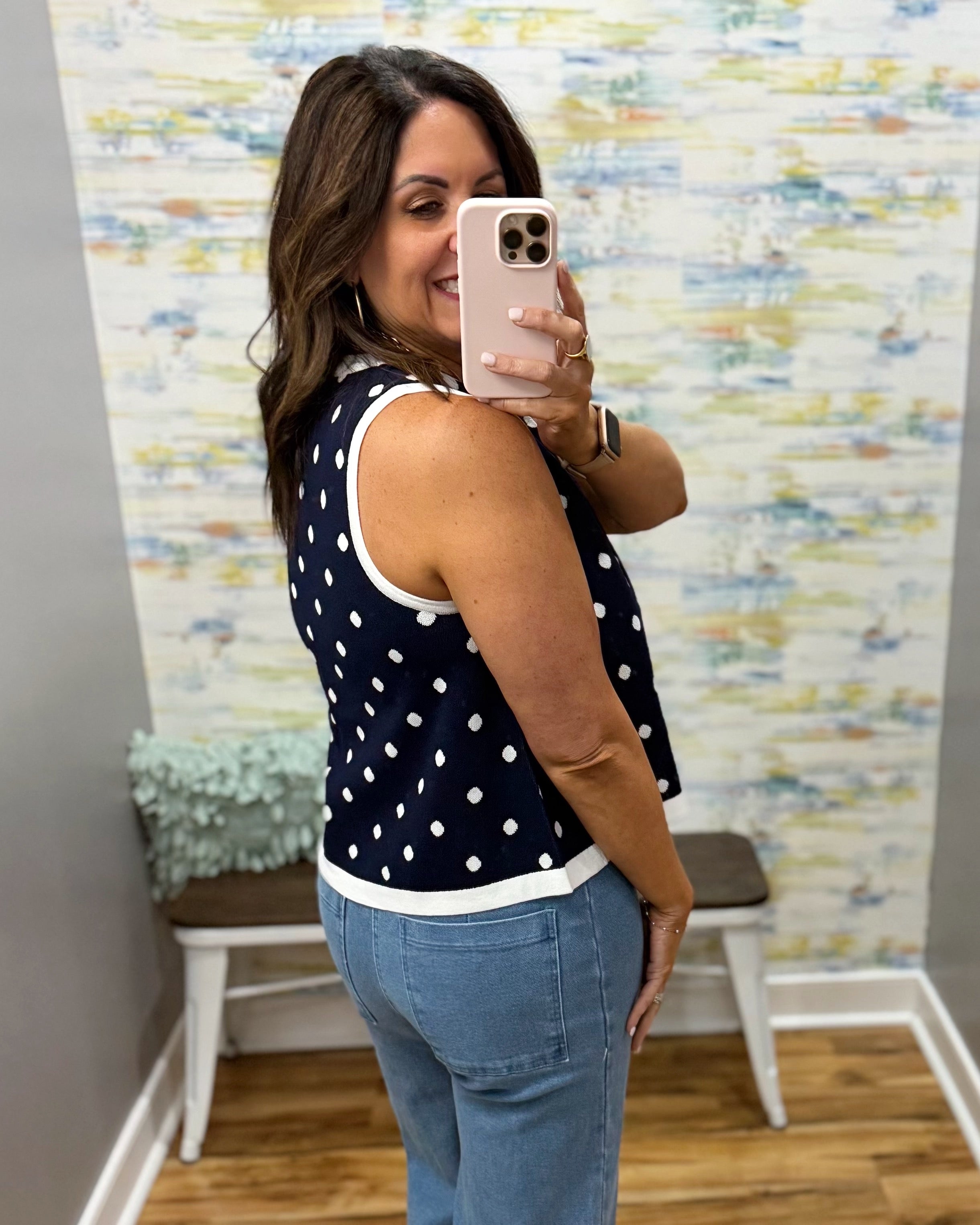 Woman taking a mirror selfie wearing a navy blue sleeveless top with white polka dots and blue jeans.