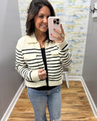 A woman takes a mirror selfie wearing a cream and black striped zip-up jacket over a black top and light blue jeans in a fitting room.