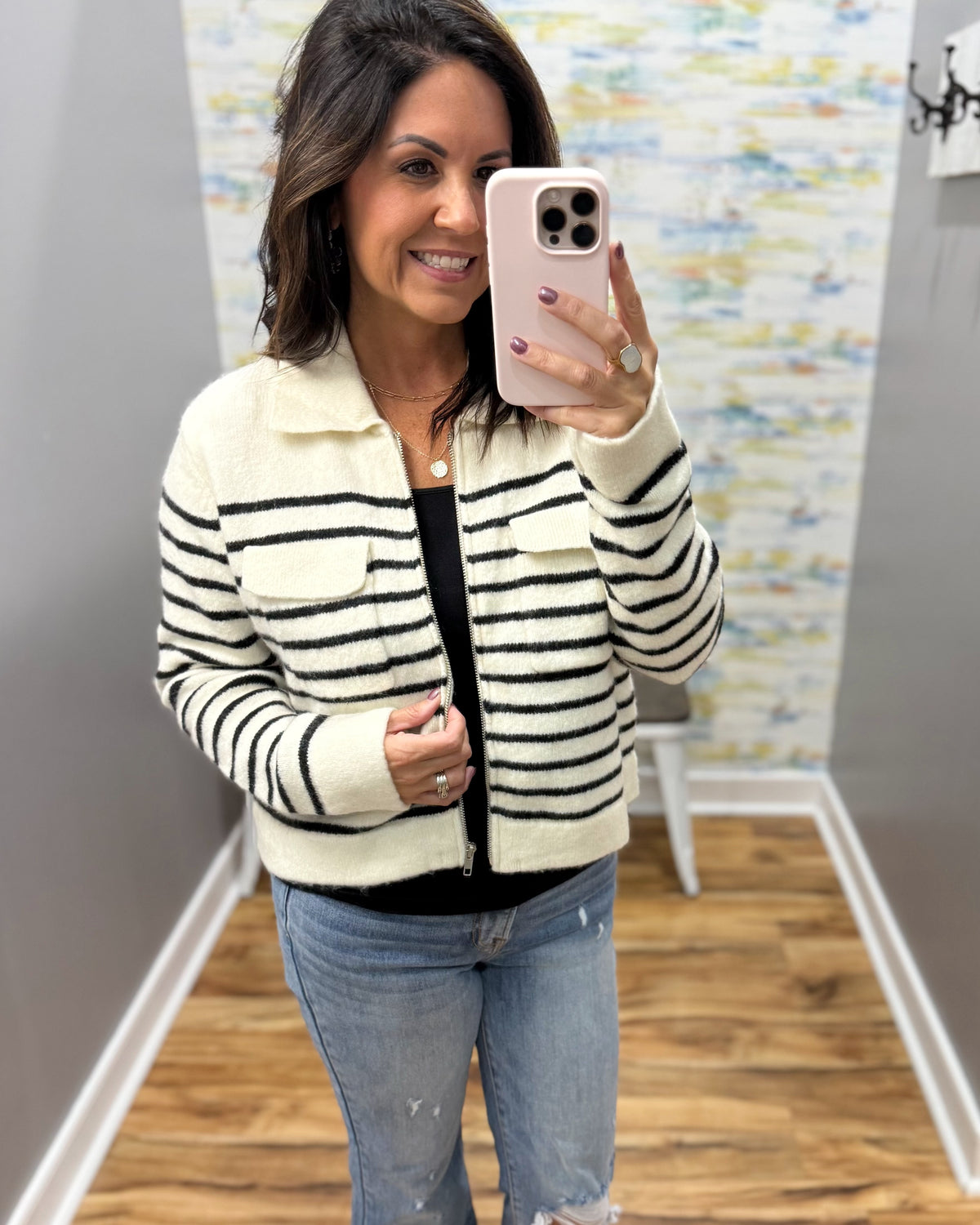 A woman takes a mirror selfie wearing a cream and black striped zip-up jacket over a black top and light blue jeans in a fitting room.