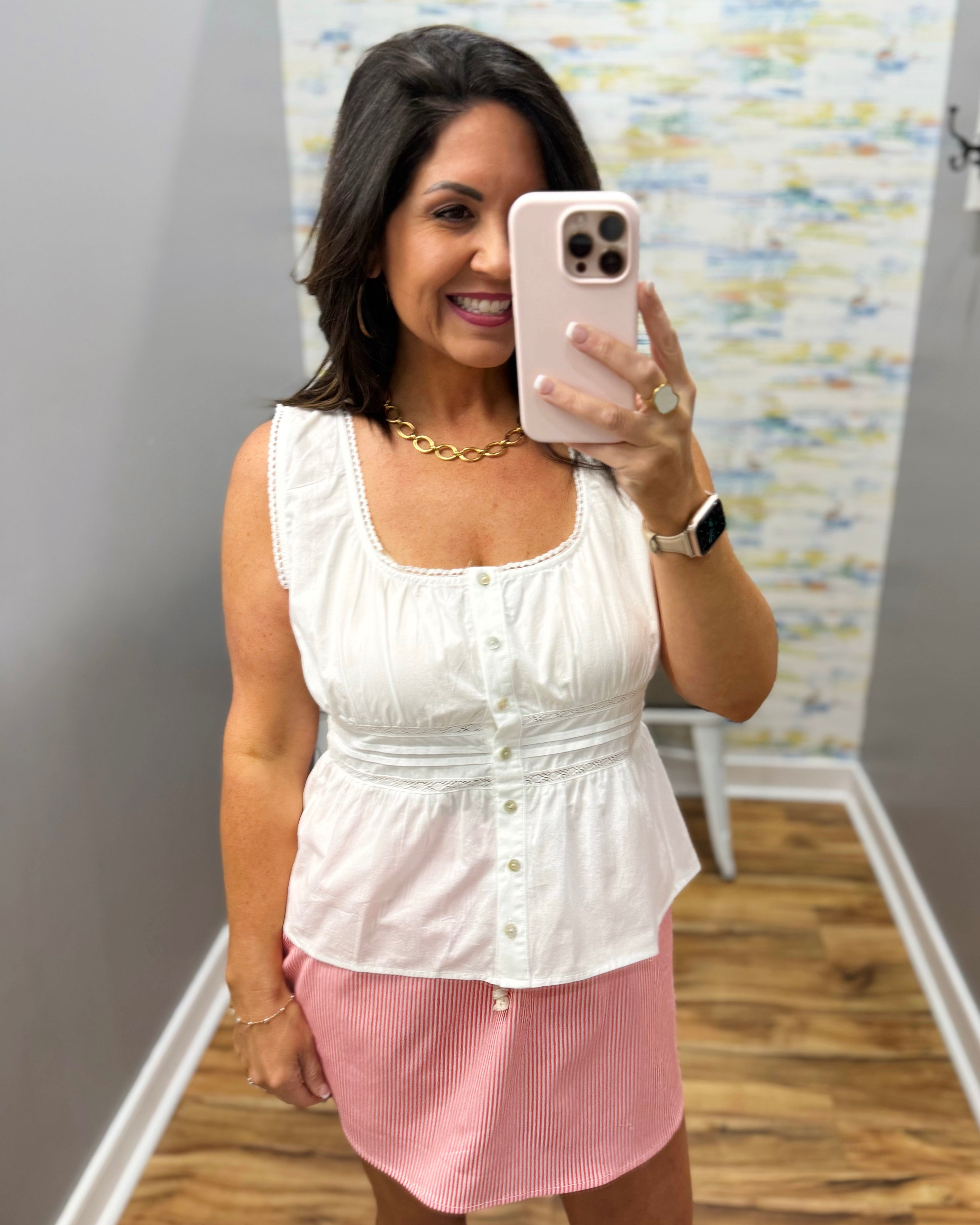 Woman taking a mirror selfie wearing a white top and pink skirt in a room with patterned wallpaper.