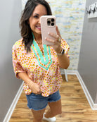Woman taking a mirror selfie wearing a colorful blouse and denim shorts in a room with a patterned wall.