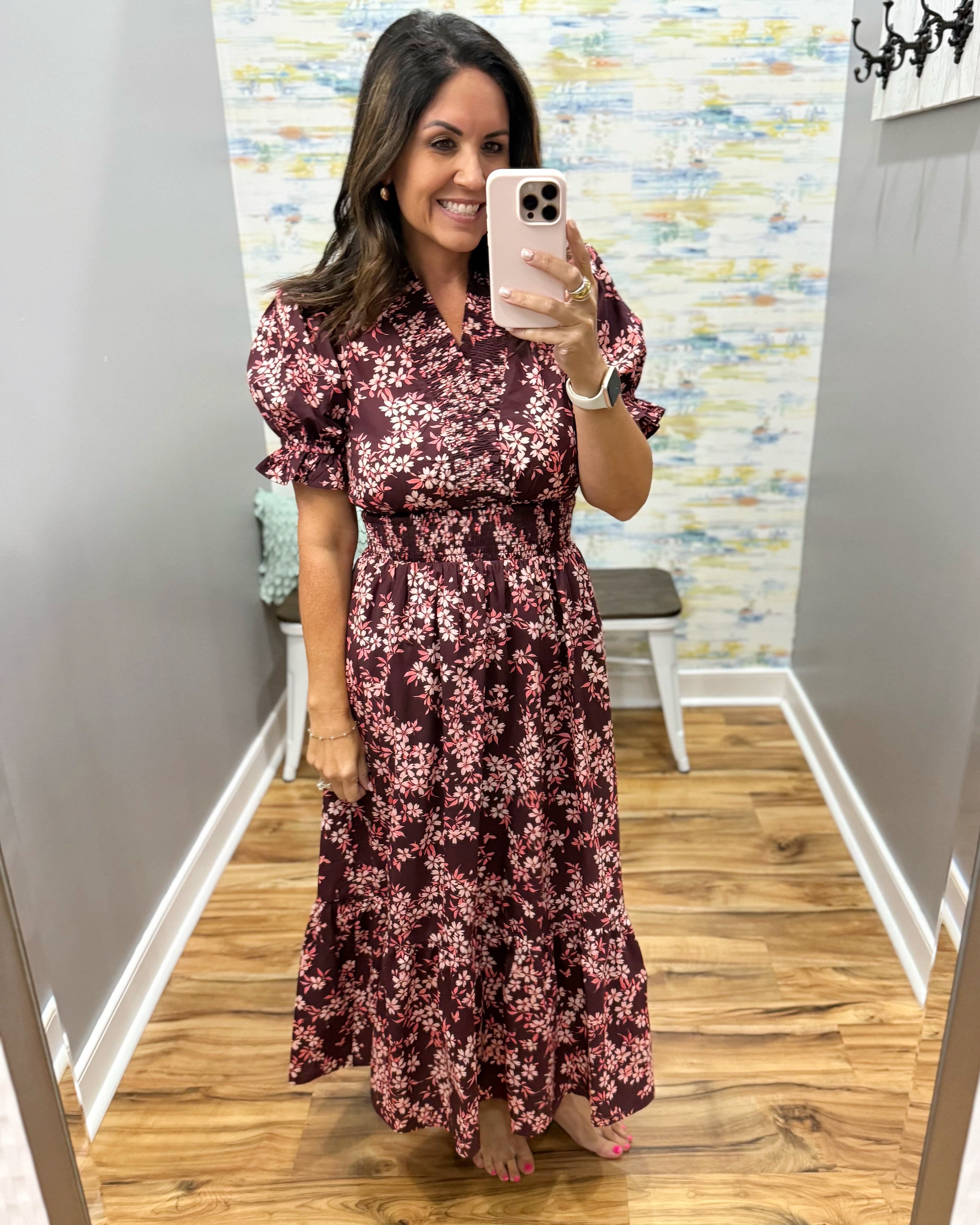 A woman takes a mirror selfie in a fitting room wearing a dark burgundy maxi dress with a pink and white floral pattern. The dress features short puff sleeves, a collared neckline, and a ruffled hem.