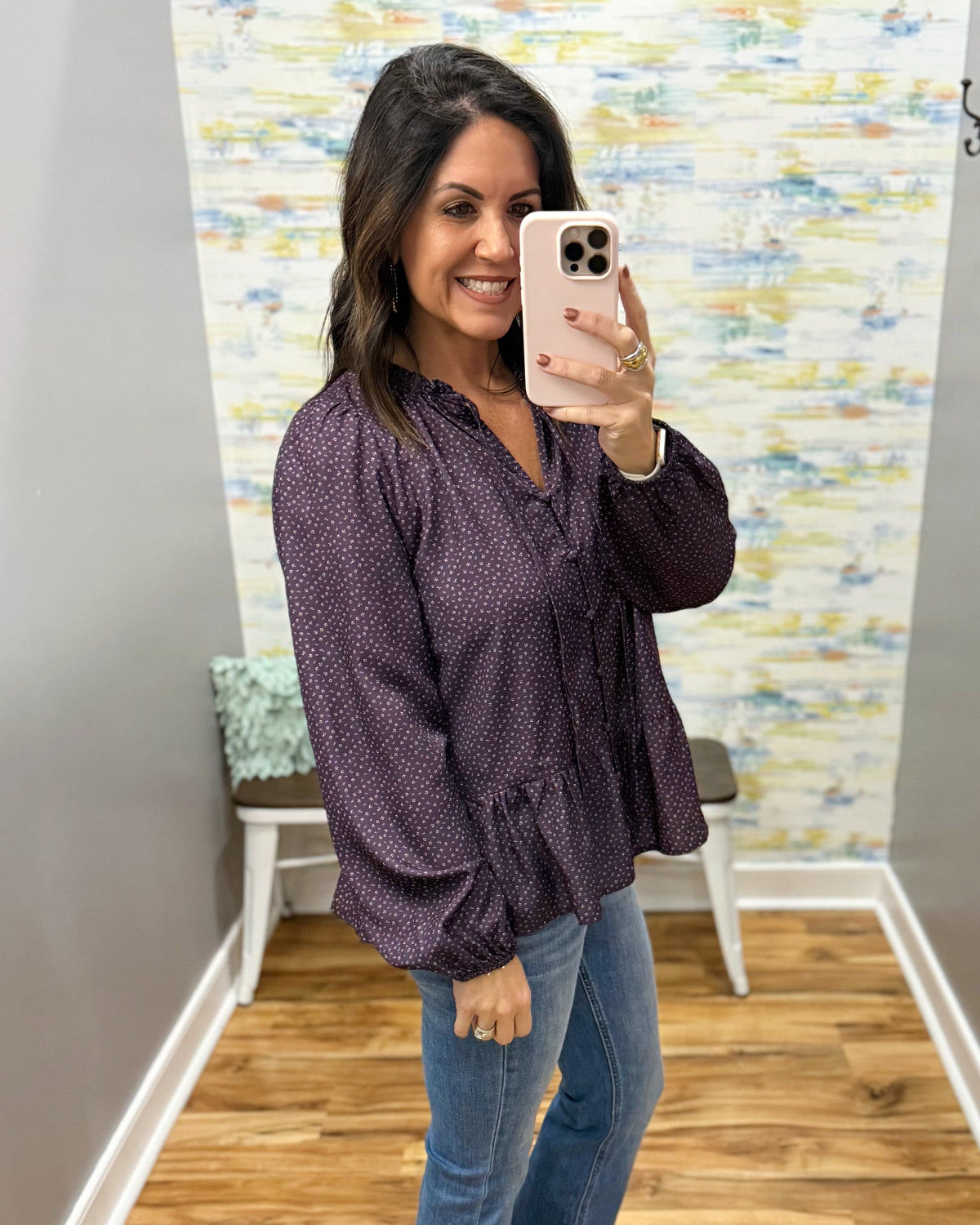 A woman takes a mirror selfie wearing a dark purple long-sleeve blouse with a small white pattern, paired with blue jeans, in front of a patterned wallpaper background.