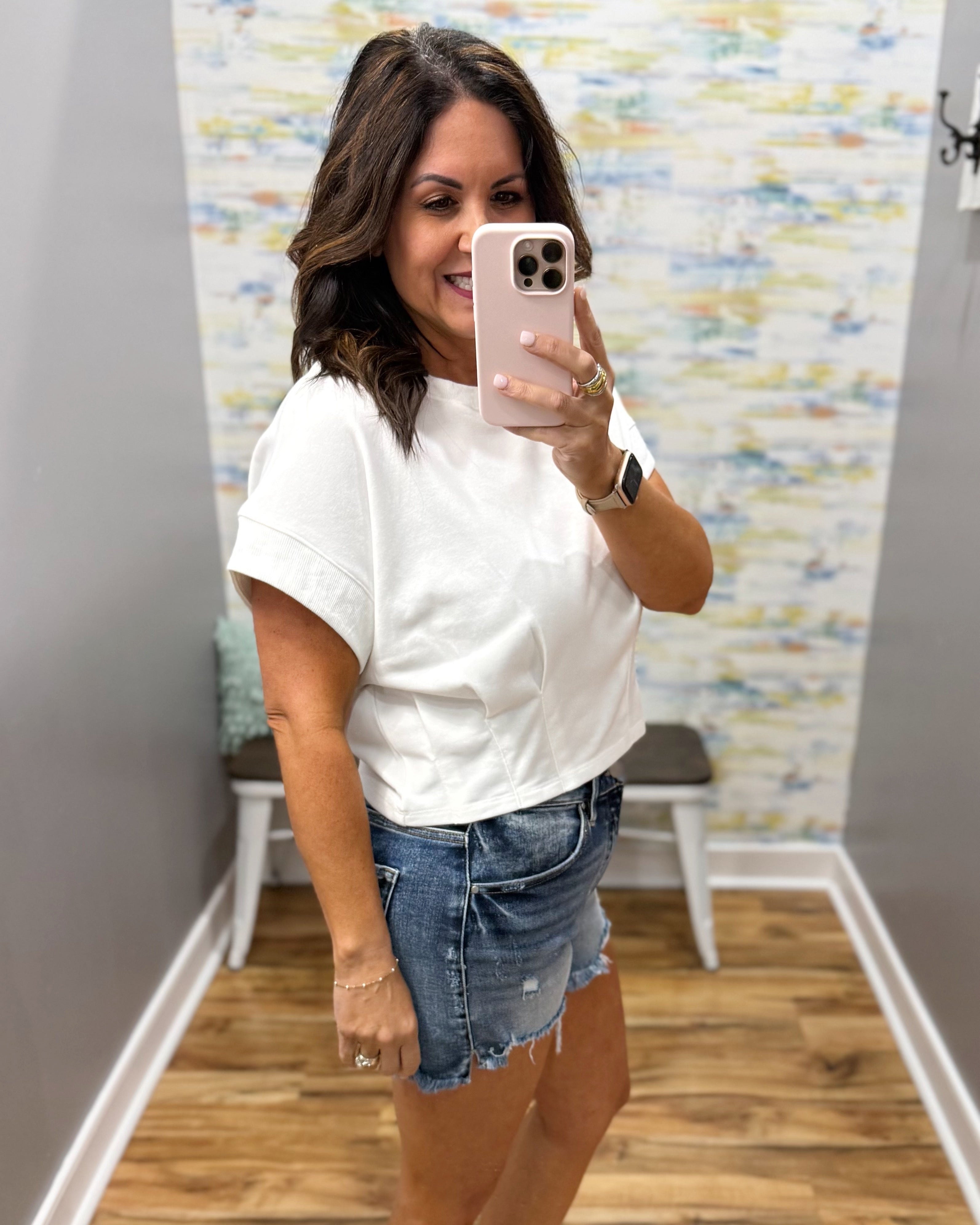 Woman taking a mirror selfie wearing a white t-shirt and denim shorts in a room with colorful wall art.