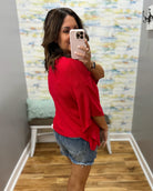 Woman taking a mirror selfie wearing a red top and denim shorts in a room with a textured wall.