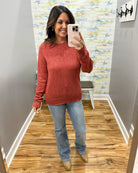 A woman takes a mirror selfie in a fitting room wearing a heathered rust-red long-sleeve knit sweater, light-wash blue jeans, and tan ankle boots.