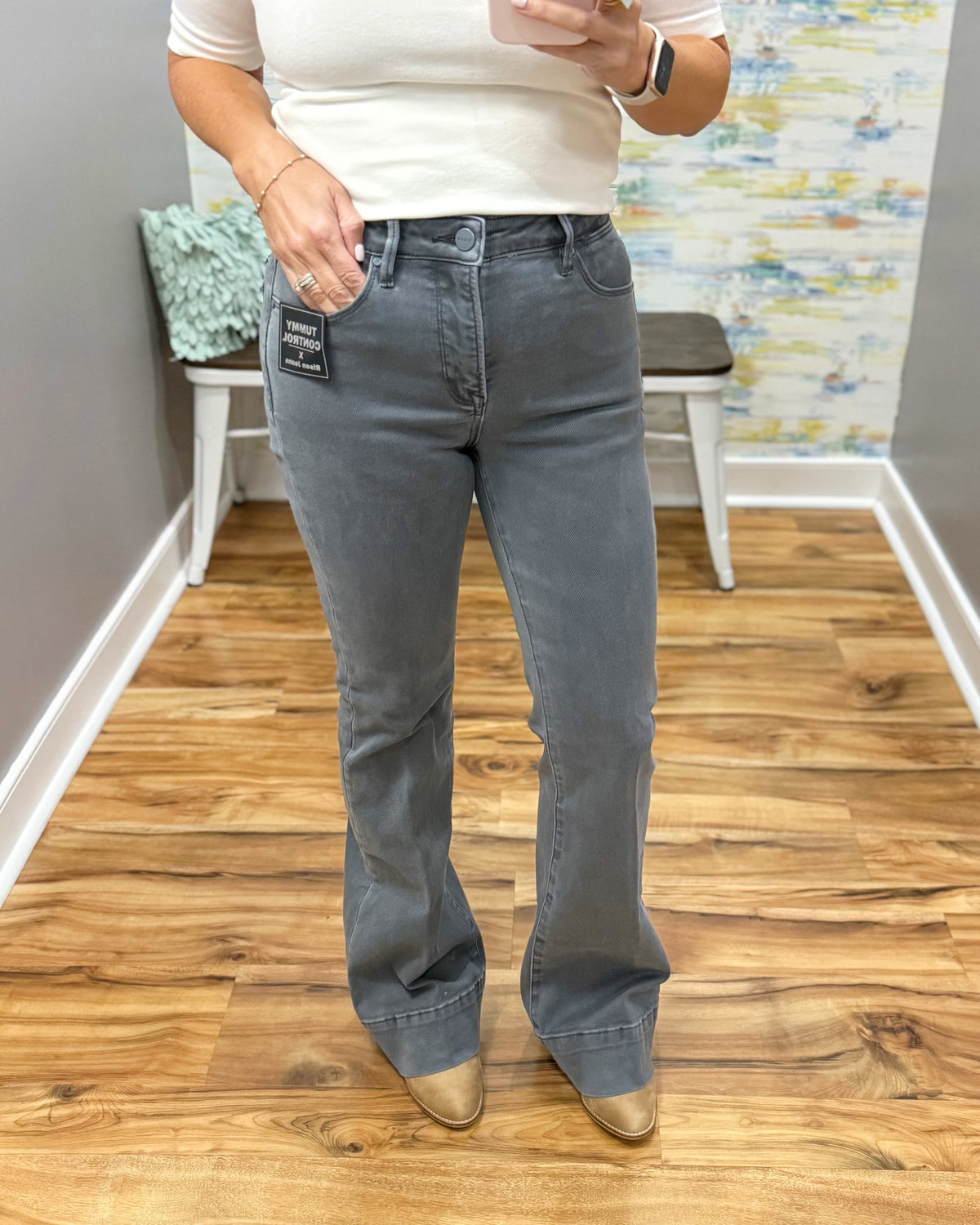 A person wearing medium-wash gray flare jeans with a white top and tan pointed-toe heels, standing on a wood floor in an indoor room.
