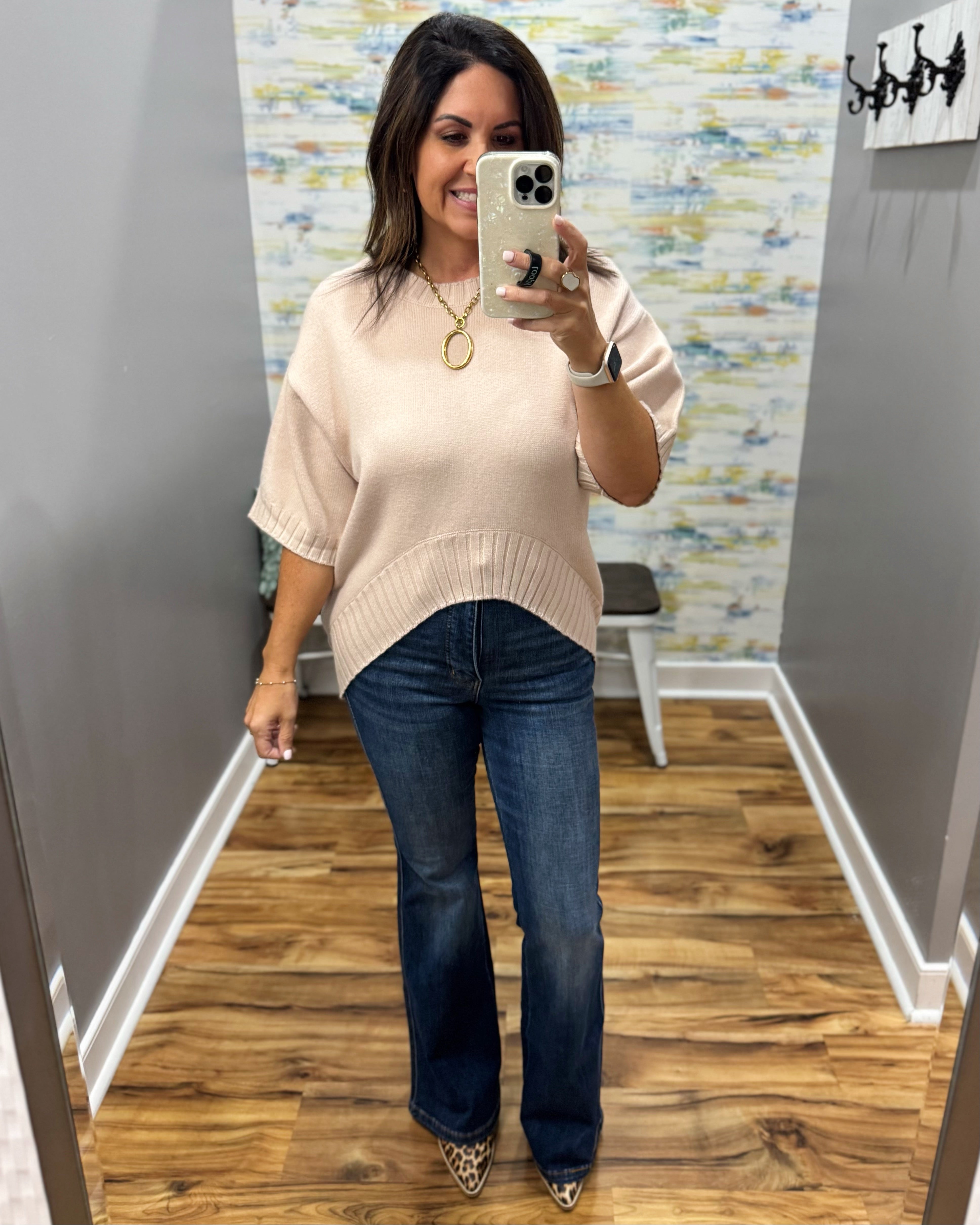 A woman takes a mirror selfie in a fitting room wearing a light beige short-sleeve knit sweater and dark blue flare jeans.