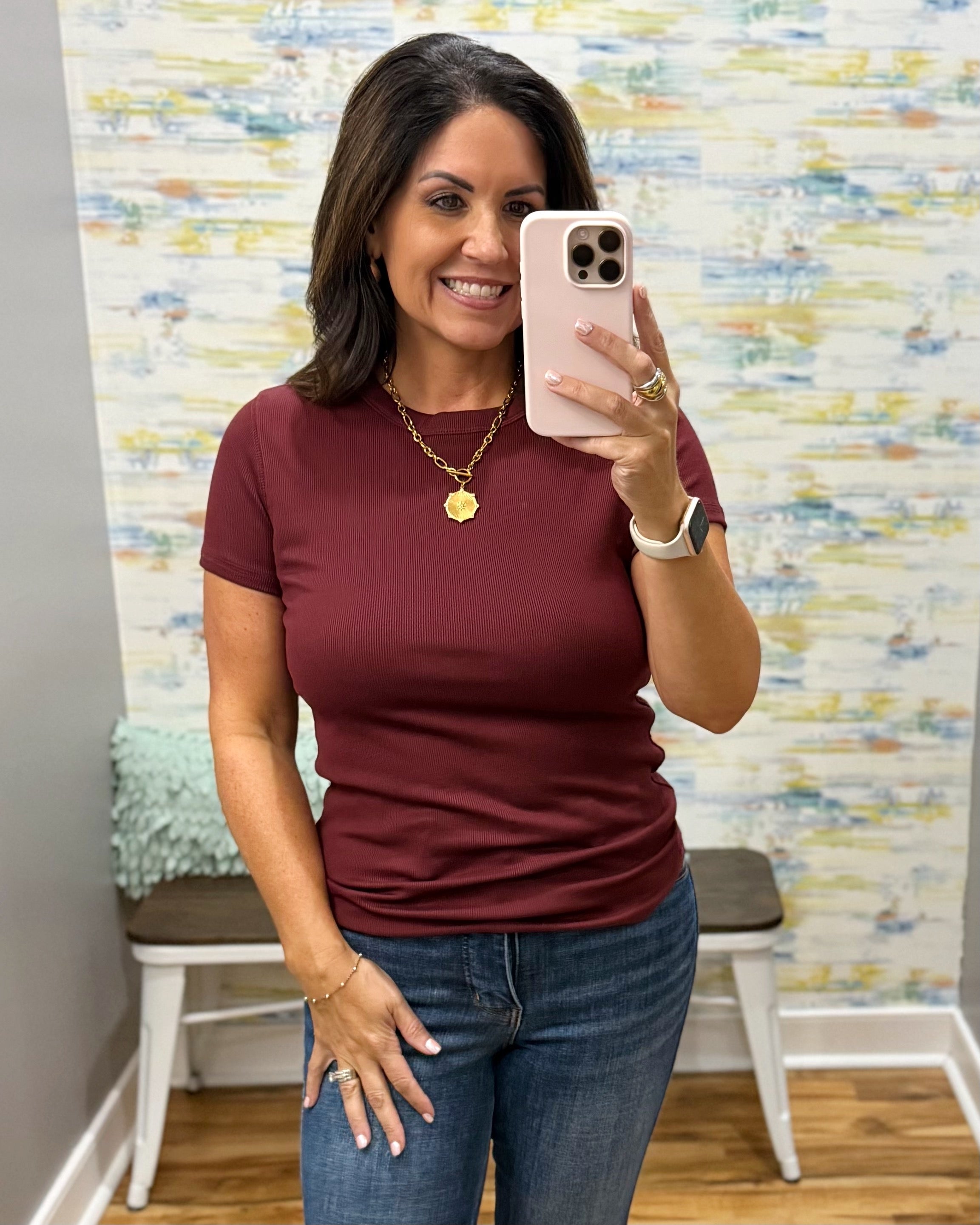 A woman taking a mirror selfie wearing a short-sleeve crewneck t-shirt in burgundy and blue jeans, standing in front of a patterned wallpaper background.