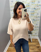 A woman takes a mirror selfie wearing a light beige short-sleeve knit top with a curved hem and blue jeans in a fitting room.