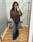 A woman takes a mirror selfie in a fitting room wearing a brown button-down shirt with rolled sleeves and blue flare jeans.