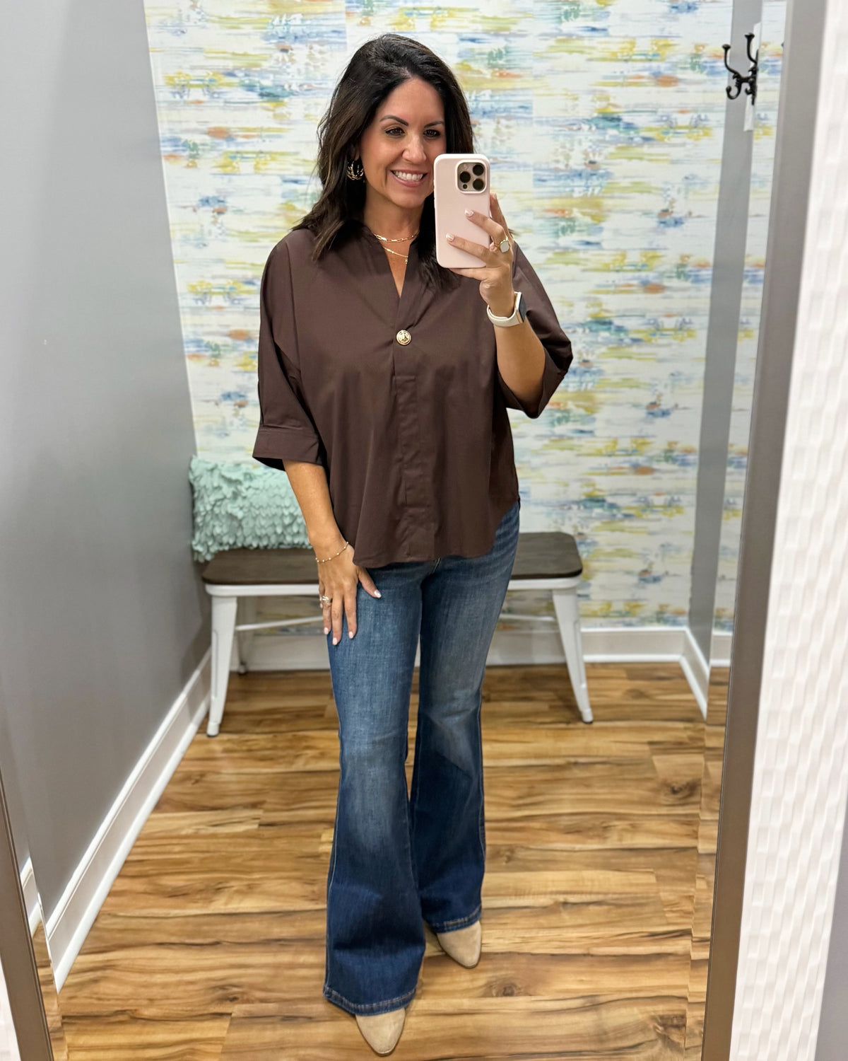 A woman takes a mirror selfie in a fitting room wearing a brown button-down shirt with rolled sleeves and blue flare jeans.