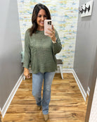 A woman takes a mirror selfie wearing a heathered sage green long-sleeve knit top, light-wash blue jeans, and tan ankle boots in a home fitting room with wood floors and abstract wall art.