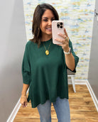 A woman takes a mirror selfie wearing a dark green, loose-fitting short-sleeve top with light blue jeans. She is wearing a gold pendant necklace and a white watch, standing in an indoor room with a wood floor and a patterned wallpaper background.