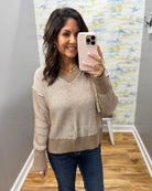 A woman takes a mirror selfie wearing a beige and brown ribbed knit sweater with dark blue jeans, holding a light pink smartphone.