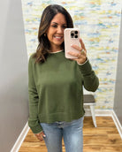 A woman takes a mirror selfie wearing an olive green long-sleeve crewneck sweater and light wash blue jeans in a fitting room with a patterned wall and wood floor.