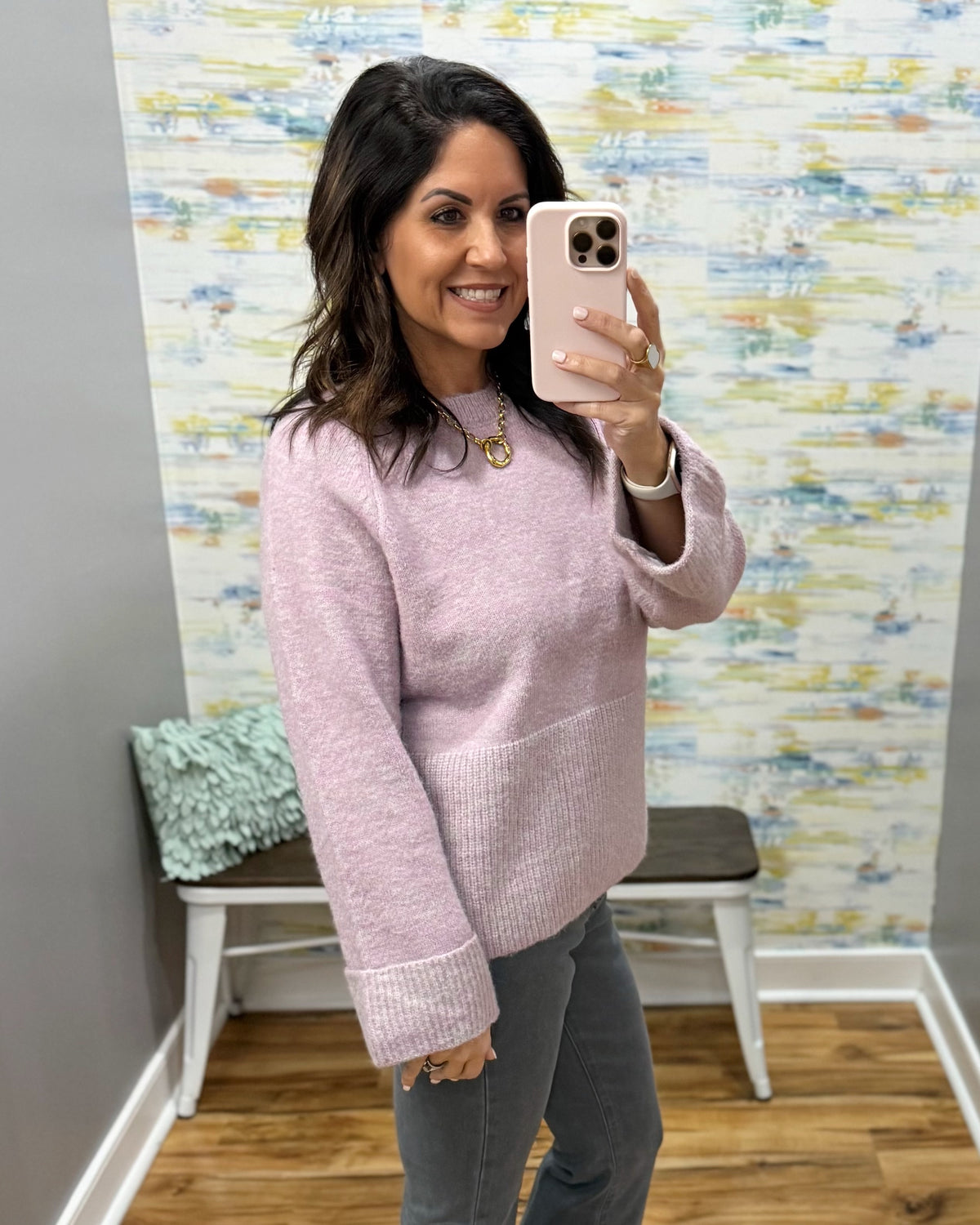 A woman takes a mirror selfie wearing a light pink knit crewneck sweater and gray pants in a home setting with patterned wallpaper and a bench in the background.