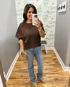 A woman takes a mirror selfie in a boutique showing a brown short-sleeve knit top and light-wash blue jeans.