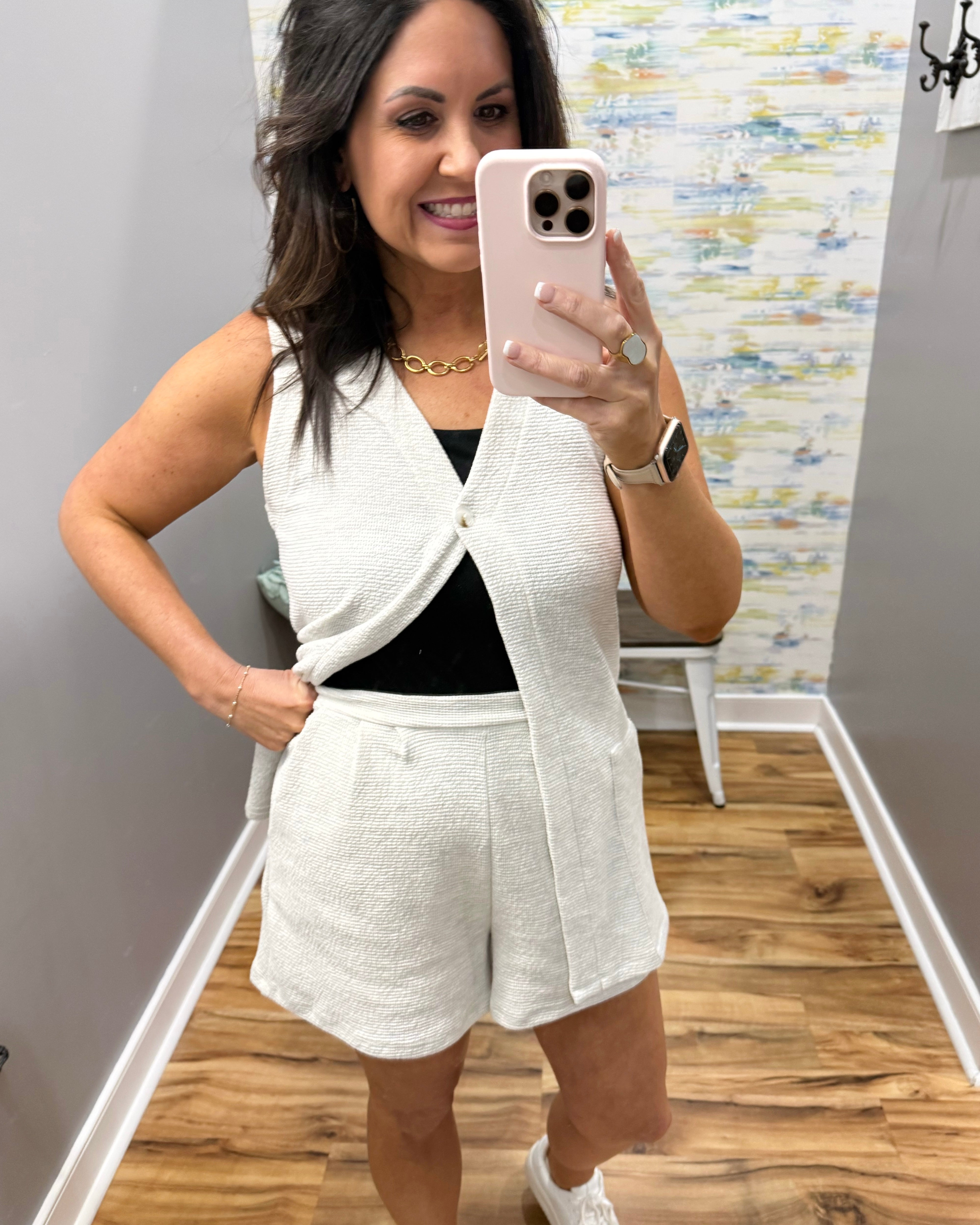Woman taking a mirror selfie wearing a white sleeveless top and shorts set.