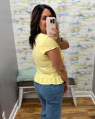 A woman taking a mirror selfie wearing a pale yellow short-sleeve peplum top and blue jeans in a room with patterned wallpaper and a bench.