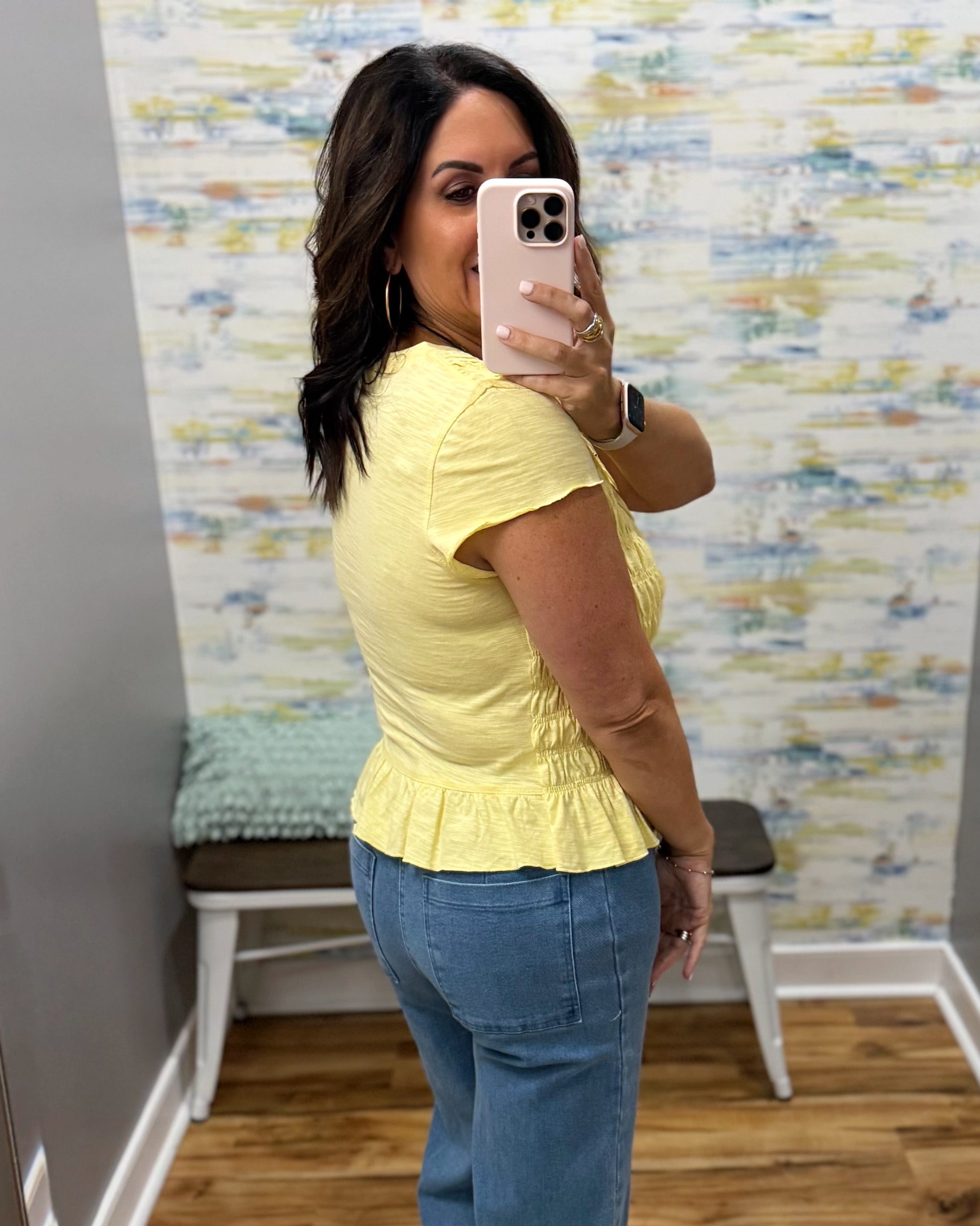 A woman taking a mirror selfie wearing a pale yellow short-sleeve peplum top and blue jeans in a room with patterned wallpaper and a bench.