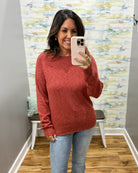 A woman takes a mirror selfie wearing a heathered rust-red long-sleeve knit sweater and light-wash blue jeans in a home fitting room with patterned wallpaper and wood floors.